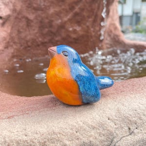 Könnte beinhalten: Keramik-Vogelfigur in Blau und Orange. Der Vogel sitzt auf einer Steinoberfläche in der Nähe eines Wasserspiels. Der Körper des Vogels ist orange, und seine Flügel und sein Kopf sind blau.