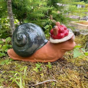 Könnte beinhalten: Keramik-Schneckenfigur mit dunkelgrünem und schwarzem Gehäuse, braunem Körper und rotem und weißem Kopfschmuck. Die Schnecke steht auf einem Bett aus grünem Moos und Gras, mit einem verschwommenen natürlichen Hintergrund.
