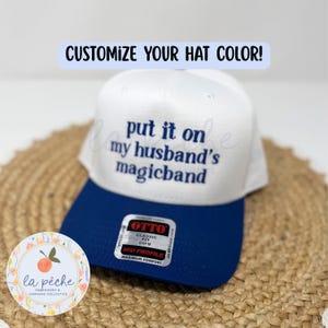 May include: A white baseball cap with a blue brim featuring the embroidered dark blue text "put it on my husband's magicband" and a sticker on the brim that reads "OTTO CLASSIC FIT OSFM MID PROFILE MAXIMUM COMFORT". A banner above reads "CUSTOMIZE YOUR HAT COLOR!"