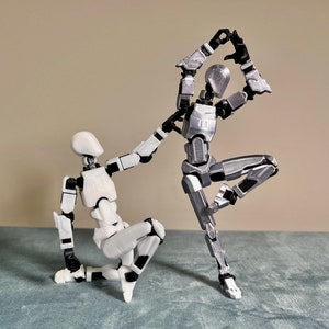 Puede incluir: Dos figuras de acción articuladas, una blanca y otra plateada, están posadas en una posición dinámica, casi acrobática. La figura blanca está de rodillas, mientras que la figura plateada está de pie sobre una pierna, con la otra pierna extendida detrás de ella.