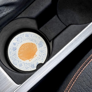 May include: A white and blue car coaster with a seashell design. The seashell is yellow and has a white outline. The coaster is in a car cup holder.