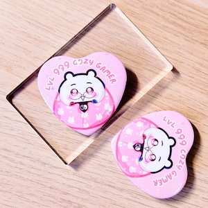 May include: Two pink heart-shaped phone grips with a cartoon bear design. The bear is wearing glasses and holding a game controller. The text "LVL 999 COZY GAMER" is printed around the edge. The grips are on a clear acrylic stand.