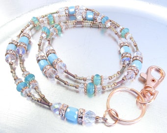 May include: A double-strand beaded lanyard with a rose gold tone metal clasp and key ring. The beads are a mix of clear crystal, light blue, and gold.