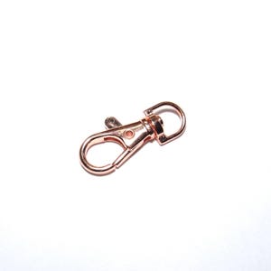 Puede incluir: Un mosquet&oacute;n giratorio de color oro rosa, con una palanca con resorte y un peque&ntilde;o anillo circular. Este cierre est&aacute; dise&ntilde;ado para sujetarse a diversos art&iacute;culos, como llaveros, bolsos o joyas. El metal tiene un acabado brillante.