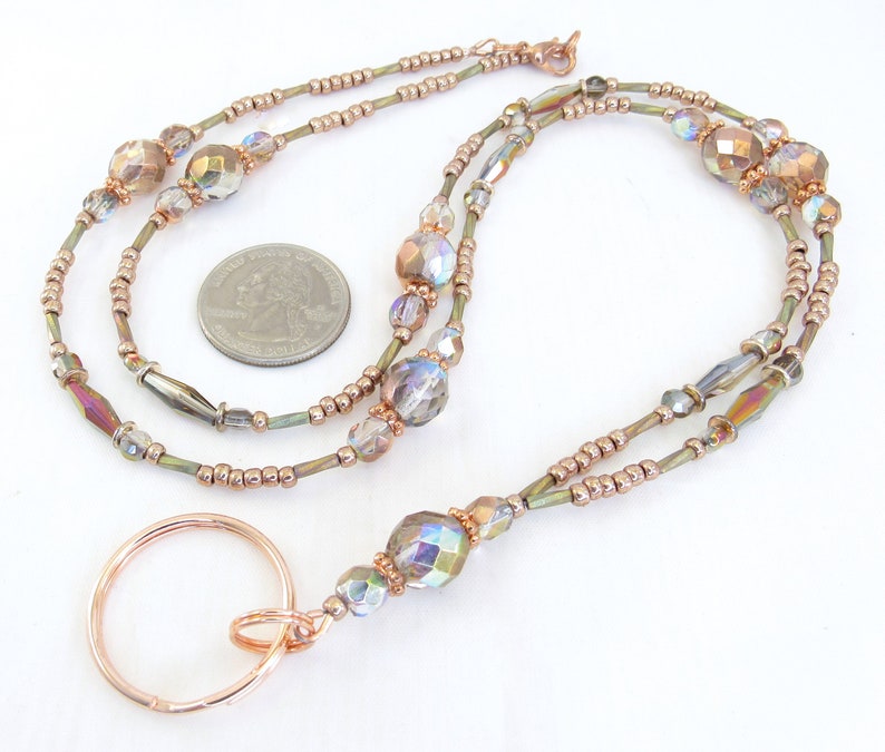 Puede incluir: Un cord&oacute;n con cuentas con un llavero de color cobre. El cord&oacute;n presenta cuentas iridiscentes y facetadas y cuentas m&aacute;s peque&ntilde;as de color cobre ensartadas en un cord&oacute;n fino de color verde oliva. El llavero es de metal color cobre.
