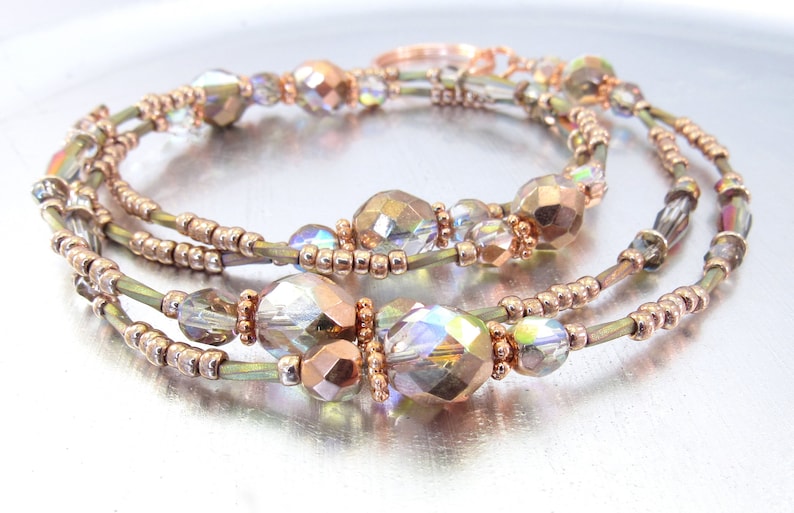 Puede incluir: Collar de m&uacute;ltiples hilos con cuentas facetadas iridiscentes y cuentas de color cobre. El collar tiene un dise&ntilde;o delicado y elegante con una mezcla de cuentas transparentes e iridiscentes, creando un efecto brillante. La joya se exhibe sobre una superficie plateada.