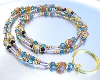 May include: A beaded lanyard with a gold key ring. The lanyard features a combination of blue, peach, and gold beads. The beads are strung together with gold-colored metal and purple-colored metal. The key ring is gold-colored.