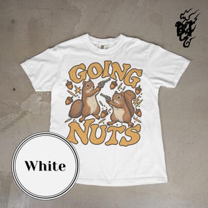 Puede incluir: Una camiseta blanca de manga corta con un gráfico de dos ardillas marrones sosteniendo revólveres. El texto "GOING NUTS" está impreso en letras grandes, negritas y de color amarillo anaranjado. Incluye bellotas y estrellas pequeñas.