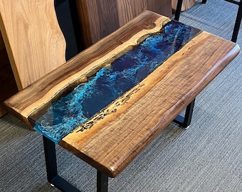 May include: A wooden coffee table with a blue epoxy river running through the center. The table has a black metal frame and is sitting on a gray carpet.