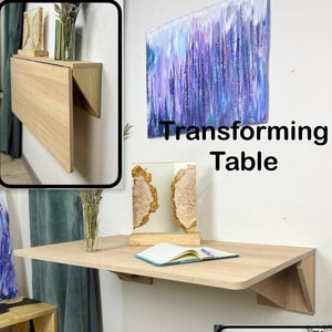 May include: A light wood folding wall table with a white wall and a painting in the background. The table is folded up and down in different positions. The text "Transforming Table" is written in black letters on the wall.