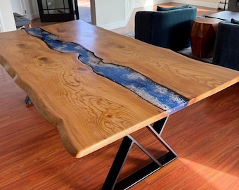 May include: A wooden dining table with a live edge top and a blue epoxy river running down the center. The table has a black metal X-shaped base.