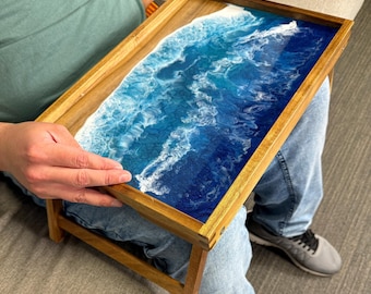 May include: Wooden serving tray with a resin ocean design. The tray features a rectangular shape with a wooden frame and legs. The resin art depicts a blue and white ocean scene, creating a coastal aesthetic.