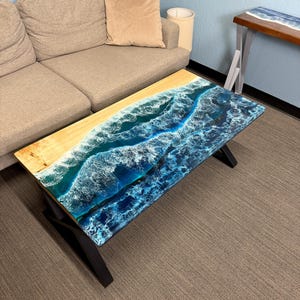 Puede incluir: Una mesa de centro rectangular con un diseño de playa. La mesa presenta una superficie de madera de color claro con un diseño de resina epoxi azul y blanco que se asemeja a las olas del océano. Se apoya en patas de metal negro.