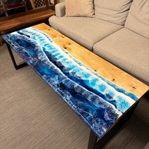 May include: A coffee table with a wooden top and black metal legs. The top features a blue and white resin design that resembles ocean waves.