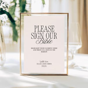 May include: A framed sign with the text "PLEASE SIGN OUR Bible" in black script. The sign also includes the text "HIGHLIGHT YOUR FAVORITE VERSE AND WRITE US A NOTE IN THE MARGIN" and "With love, ELLIE AND CHRIS 10.11.26". The frame is gold.