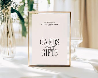 May include: A framed sign with gold trim displays the text "THE WEDDING OF ELLIE AND CHRIS 10.11.26" at the top, with "CARDS and GIFTS" in elegant script below. The sign is set on a white surface.