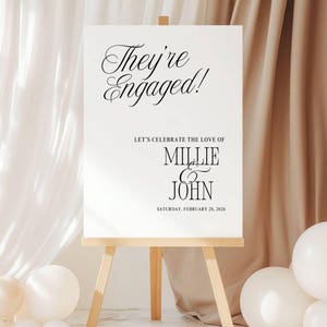 May include: A white sign on a wooden easel with the text "They're Engaged!" in elegant black script. Below, it reads "Let's Celebrate the Love of Millie & John" and the date "Saturday, February 28, 2026".