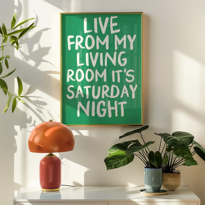 May include: A green framed poster with white text that reads "Live from my living room it's Saturday night". The poster is hanging on a white wall with a brown lamp and two green plants in front of it.