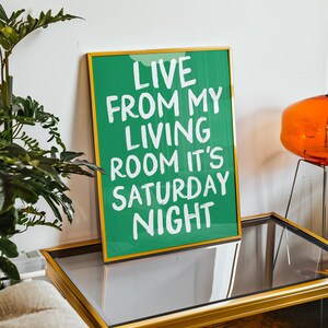 May include: A green framed print with white text that reads "Live from my living room it's Saturday night". The print is sitting on a gold-framed glass table.
