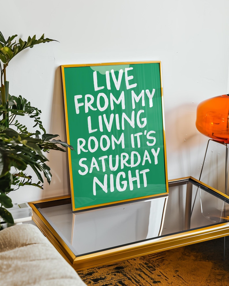 May include: A green framed print with white text that reads "Live from my living room it's Saturday night". The print is sitting on a gold-framed glass table.