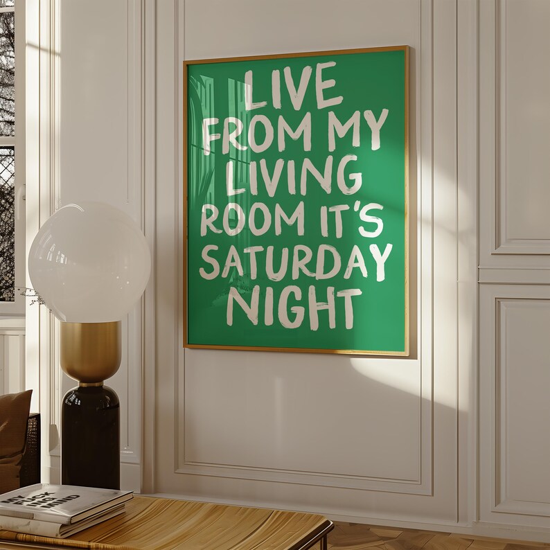 May include: A green poster with white text that reads "Live from my living room it's Saturday night". The poster is framed in gold and hanging on a white wall.