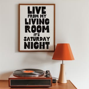 May include: A framed print with the text "Live from my living room it's Saturday night" in black lettering on a white background. A vintage-style record player and a red lamp are on a wooden surface below the print.