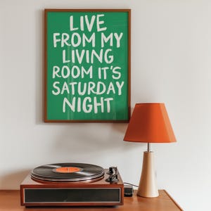 May include: A framed green poster with white text that reads "Live from my living room it's Saturday night". A record player with a black vinyl record on the turntable is on a wooden surface. A red lamp with a wooden base is on the right side of the image.