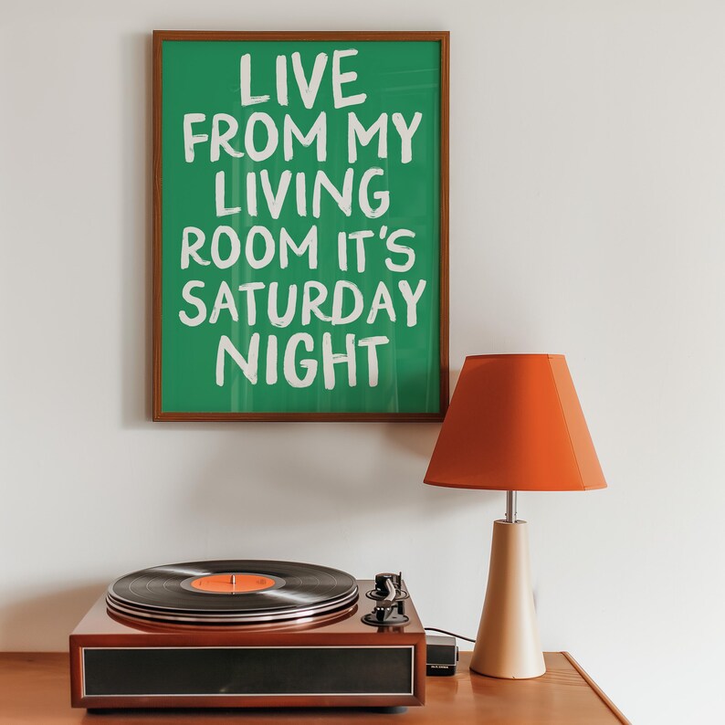 May include: A framed green poster with white text that reads "Live from my living room it's Saturday night". A record player with a black vinyl record on the turntable is on a wooden surface. A red lamp with a wooden base is on the right side of the image.