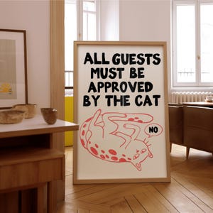 May include: A framed print with the text "ALL GUESTS MUST BE APPROVED BY THE CAT." The print features a red cartoon cat with spots, lying on its back. A speech bubble says "NO."