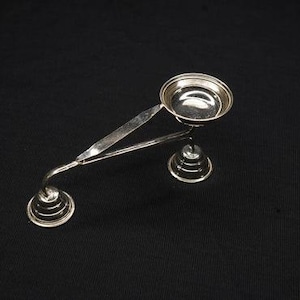May include: A silver-colored, ornate incense burner with a small bowl and two spiral-shaped bases. The burner has a long, thin handle and is set against a black background. The bowl is round and has a reflective surface.