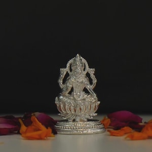 May include: A metallic silver-colored figurine depicting a deity seated in a cross-legged position upon a lotus flower base. The figure features four arms, an ornate crown, and detailed traditional attire, all rendered with a polished finish.