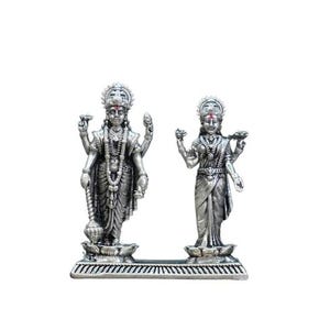 May include: A silver-colored statue depicting two figures, likely deities, standing on a decorative base. The figures are adorned with detailed jewelry and headdresses. The statue is set against a plain white background.