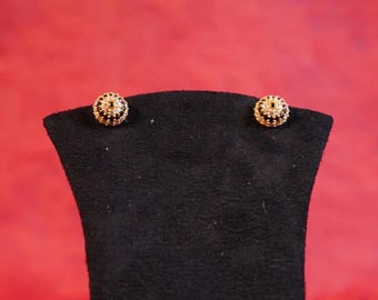 May include: A pair of gold-toned stud earrings with a black and white design. The earrings are displayed on a black jewelry stand against a red background. The earrings have a circular shape with intricate detailing.
