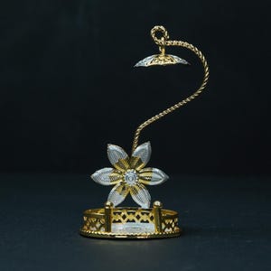 May include: A decorative, gold and silver-toned metal flower-shaped object. The flower sits atop a curved, twisted stem, with a small, matching shade at the top. The base is a circular, openwork design. The object is set against a dark background.