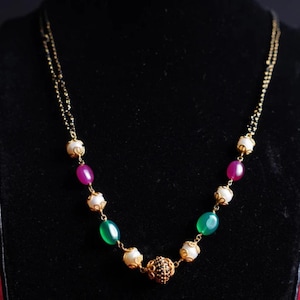 Puede incluir: Un collar dorado con una cadena delicada. El collar presenta cuentas ovaladas blancas, verdes y magenta alternadas, intercaladas con una cuenta dorada central ornamentada. El collar se muestra sobre un fondo negro.