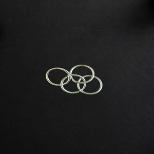 May include: Silver interlocking rings arranged in a cluster, set against a black background. The rings are smooth and shiny, with a minimalist design. The image is well-lit, highlighting the metallic texture and shape of the rings.
