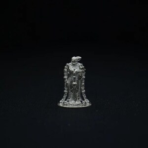 May include: A small, silver-colored figurine of a robed figure with ornate detailing. The figure has a crown and holds an object in its hand. The figurine is standing on a circular base against a black background.