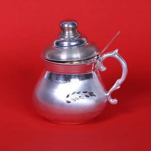 May include: A silver-colored sugar bowl with a lid and handle. The bowl has a rounded shape with a decorative leaf design. A small spoon rests on the rim. The background is a solid red.