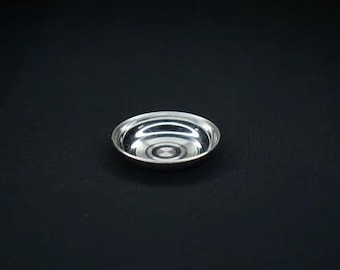 May include: A small, shiny silver bowl with a circular design in the center. The bowl is set against a black background, highlighting its reflective surface. This decorative bowl is suitable for serving small portions.
