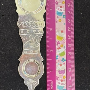 May include: A silver-toned metal object with a decorative engraved pattern, featuring two circular indentations and a scalloped base. A pink Westcott ruler is positioned alongside, showing the object measures approximately 5 inches in length.