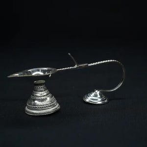May include: A silver-colored inkwell set with a decorative design. The set includes a small, cone-shaped inkwell and a pen holder with a curved arm and a small dish for the pen. The inkwell is approximately 2 inches tall.