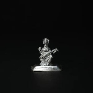 May include: A silver figurine of a deity playing a stringed instrument, set against a black background. The figure is detailed, with intricate carvings and a serene expression. The figurine is mounted on a rectangular base.