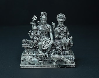 Puede incluir: Una detallada escultura de color plateado que representa deidades hindúes. Las figuras centrales están sentadas, con figuras y animales adicionales, incluyendo un pavo real y un toro, en la base. La escultura está sobre un fondo oscuro.