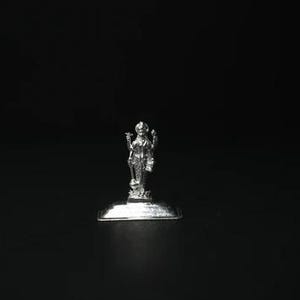 May include: A small, silver-colored statue of a deity, standing on a rectangular base. The figure is highly detailed, with intricate clothing and ornamentation. The statue is set against a black background.