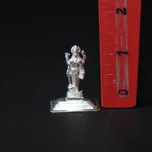 May include: A small, silver-colored statue of a deity, standing on a rectangular base. The statue is approximately 1 inch tall, with intricate details visible. A red ruler is positioned next to the statue for scale, against a black background.