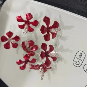 May include: Five red flower-shaped earrings with silver accents. The earrings are displayed on a white surface. Each flower has five petals and a cluster of silver stones in the centre.