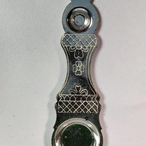 May include: A silver-toned metal object with a long, slender, contoured body featuring engraved floral and cross-hatched patterns. It has a rounded, recessed circular basin at the bottom and a smaller, circular indentation near the top finial.