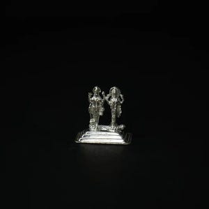 May include: A silver-colored statue depicting two figures standing side-by-side on a rectangular base. The figures are detailed with intricate designs and stand against a black background. The statue appears to be made of metal.
