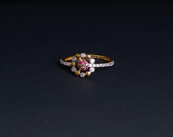 May include: A gold ring with a floral design. The center features a cluster of small red and green gemstones surrounded by white and gold accents. The band is lined with small, clear stones.