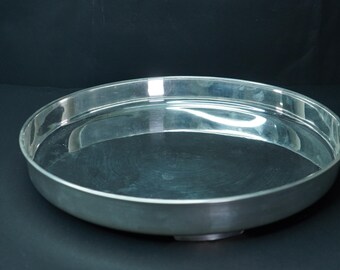 May include: A round, silver-colored metal serving tray with a wide, shallow bowl. The tray has a reflective surface and a simple, elegant design. The tray is suitable for serving food or displaying decorative items.